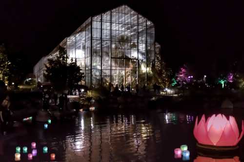 A glass greenhouse illuminated at night, surrounded by colorful lights and a pond with floating lanterns and a lotus flower.