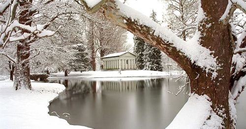 A serene winter scene featuring a snow-covered landscape, a pond, and a classical building in the background.