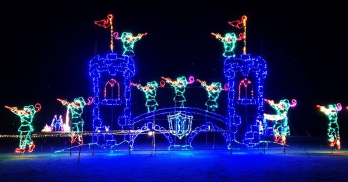 Colorful holiday lights form a castle with figures in green, celebrating with flags and a bridge in a nighttime setting.