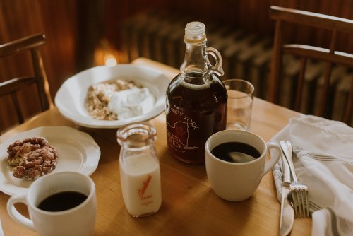 A cozy breakfast table with coffee, milk, maple syrup, granola, and a cookie on a wooden surface.