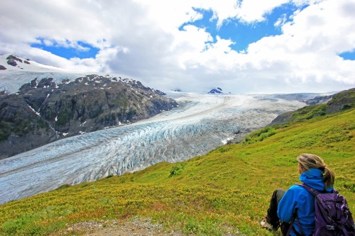 A person sits on a grassy hillside, gazing at a vast glacier surrounded by mountains and a cloudy sky.