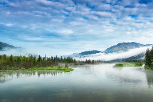 A serene river flows through a misty landscape, surrounded by lush trees and mountains under a cloudy sky.