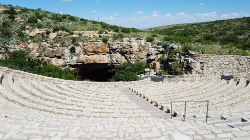 An outdoor amphitheater with stone seating, surrounded by greenery and a rocky cave entrance in the background.