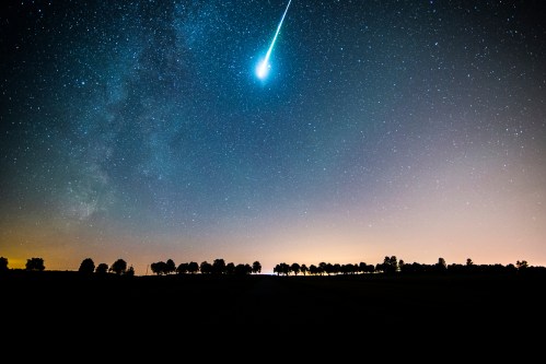 A bright meteor streaks across a starry night sky, with silhouettes of trees against a glowing horizon.