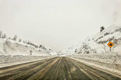 A snowy road flanked by high snowbanks, with a caution sign indicating a curve ahead. Visibility is low due to snowfall.