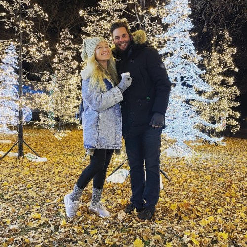 A couple smiles together in a festive outdoor setting with illuminated trees and fallen leaves.