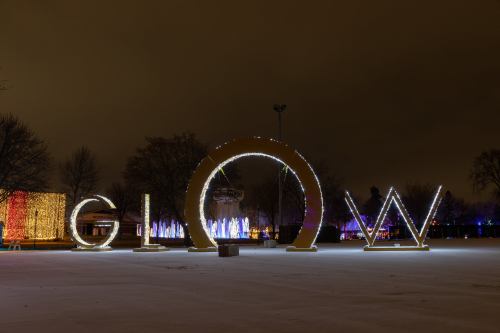 Illuminated "GLOW" sign in a snowy park at night, surrounded by colorful light displays and trees.