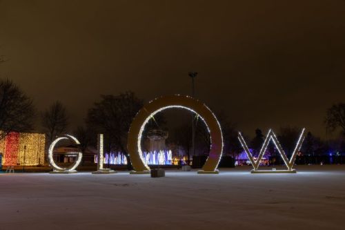 A large illuminated sign spelling "GLOW" in a snowy park, surrounded by colorful lights and trees at night.