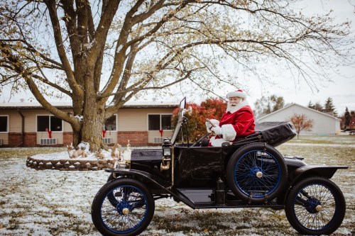 Santa Claus in a vintage car, smiling and waving, with a snowy landscape and a tree in the background.