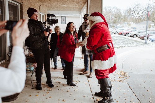 A woman in a red sweater hands a gift to Santa, surrounded by onlookers and a camera crew on a porch.