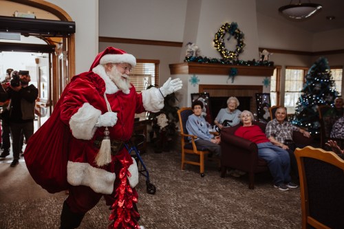 Santa Claus joyfully enters a room filled with seated seniors, spreading holiday cheer in a festive setting.