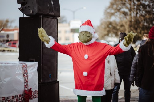 A person in a Grinch costume wearing a Santa hat and red outfit, standing with arms outstretched at a festive event.