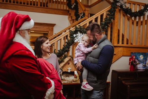 A family interacts with Santa Claus in a festive home, with a child being held and smiling at her father.