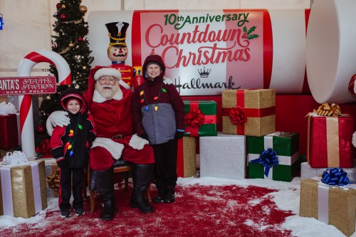 Santa Claus sits with two children in festive attire, surrounded by Christmas decorations and gifts.