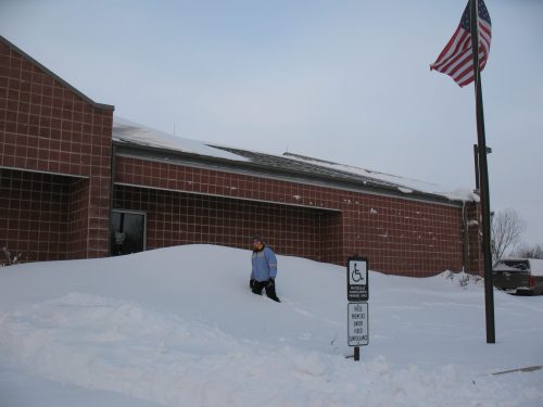 A person in winter clothing stands on a large snowbank near a brick building and an American flag.