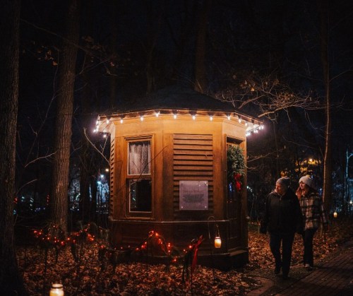 A cozy wooden cabin adorned with lights, surrounded by autumn leaves, as two people stroll by at night.