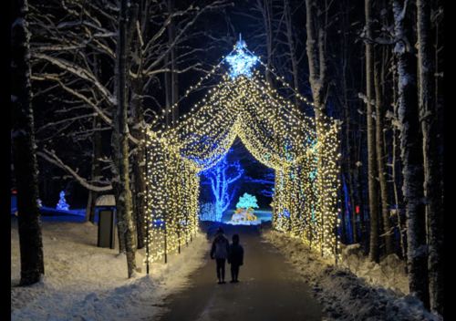 A snowy path lit by festive lights, with two people walking under a glowing archway in a winter wonderland.