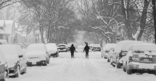 Two people walk down a snow-covered street lined with parked cars, surrounded by trees in a winter scene.