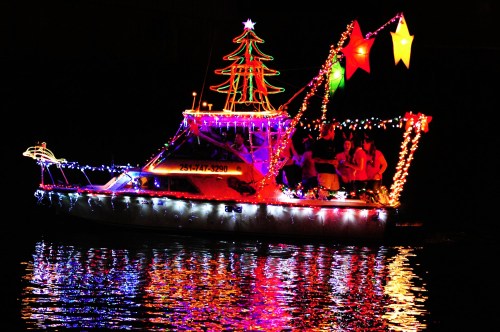 A brightly lit boat decorated with Christmas lights and a tree, reflecting on dark water at night.