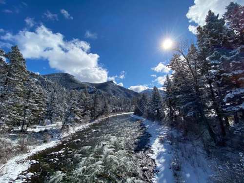 A snowy river winds through a forested landscape under a bright blue sky with fluffy clouds and a shining sun.