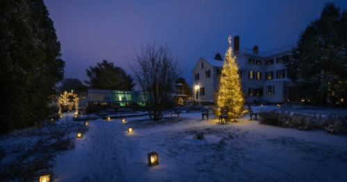 A snowy landscape at dusk featuring a lit Christmas tree and glowing lanterns in a garden near a house.