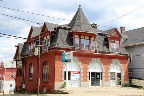 Historic red brick building with arched windows, a steep roof, and a "For Sale" sign in a small town setting.