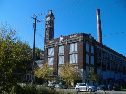 Historic brick factory building with a clock tower and smokestack, surrounded by trees and power lines under a clear blue sky.