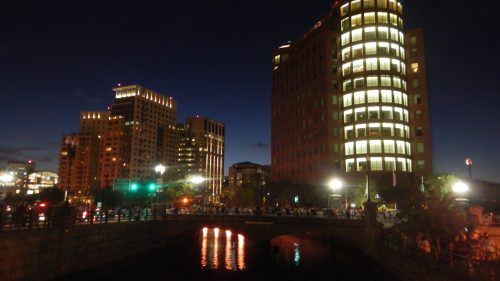 City skyline at night with illuminated buildings and a bridge over a calm waterway.