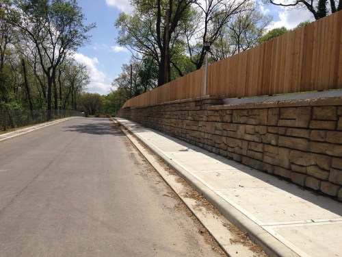 A paved road lined with a stone wall and wooden fence, surrounded by trees under a blue sky.