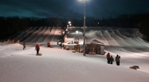 A snowy hill at night with people sledding and a small building, illuminated by lights.
