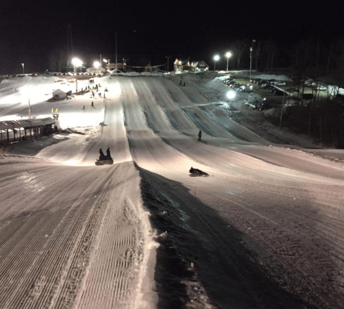 Nighttime view of a snow tubing hill with people sliding down and lights illuminating the snowy landscape.