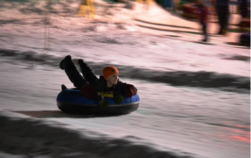 A person in a black and blue snow tube slides down a snowy slope, smiling and wearing an orange hat.