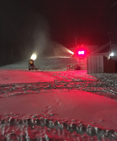 Snow-covered landscape at night with a snow cannon and red lights illuminating the scene.