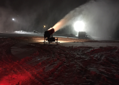 A snowmaking machine operates at night, spraying snow in a snowy landscape illuminated by bright lights.