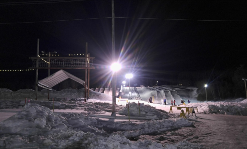 Nighttime scene of a snowy landscape with ski slopes, illuminated by bright lights and a ski lift structure.