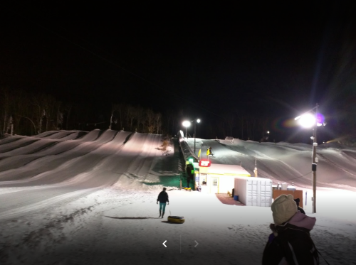 Nighttime view of a snowy slope with illuminated paths and a small building at the base. People are enjoying the winter scene.