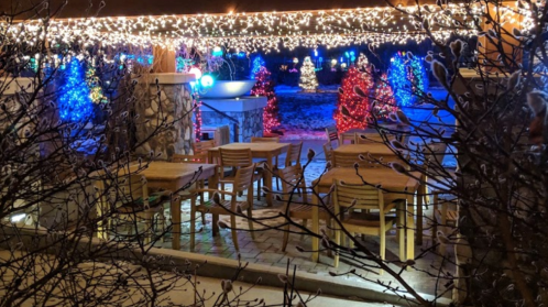 A cozy outdoor dining area adorned with colorful holiday lights and decorated trees in a snowy setting.