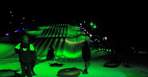 A snowy hill illuminated in green lights, with people tubing and enjoying the night.