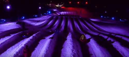 A snowy hill with colorful lights, featuring people sledding down the slopes at night.