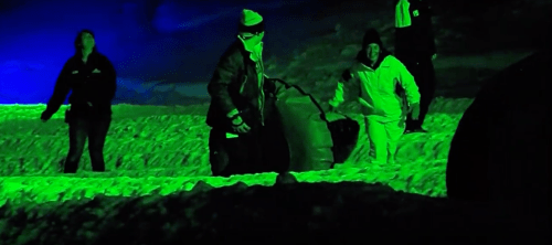 Two people in winter clothing work in a snowy landscape illuminated by green light, with a tire nearby.