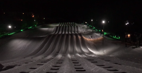 A snowy tubing hill illuminated at night, featuring multiple lanes and green lights along the sides.