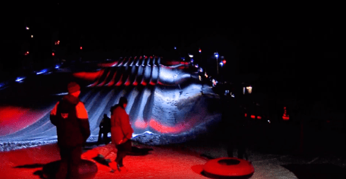 A snowy tubing hill illuminated by colorful lights at night, with people walking and preparing to slide down.