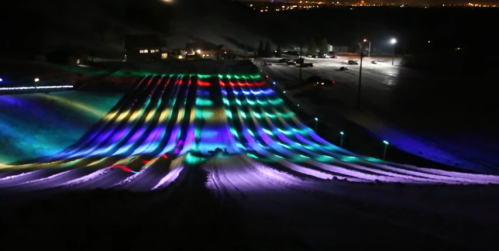 A brightly lit snow slope at night, featuring colorful lights creating vibrant patterns across the snowy surface.