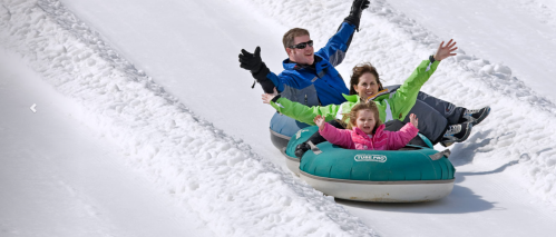 A family joyfully sleds down a snowy hill on inflatable tubes, with arms raised in excitement.