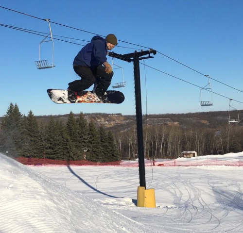A snowboarder jumps above a ski lift line on a snowy slope, with trees and a clear blue sky in the background.