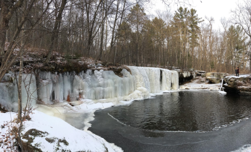 A frozen waterfall surrounded by snow and trees, with a calm pool of water at the base.