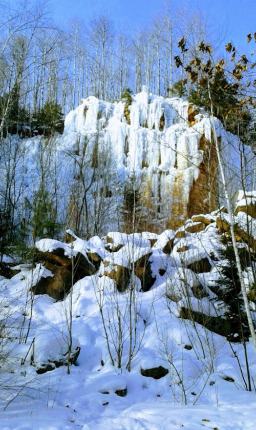 A snowy landscape featuring a frozen waterfall cascading down a rocky cliff, surrounded by bare trees.