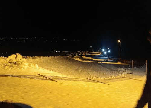 A snowy landscape at night, illuminated by distant lights and street lamps, with a clear sky above.