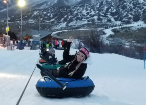 A woman in a winter hat waves while sitting on a snow tube, with a snowy landscape and people in the background.