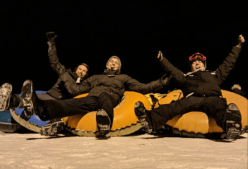 Three people joyfully sitting on inflatable tubes, smiling and raising their arms in a snowy nighttime setting.